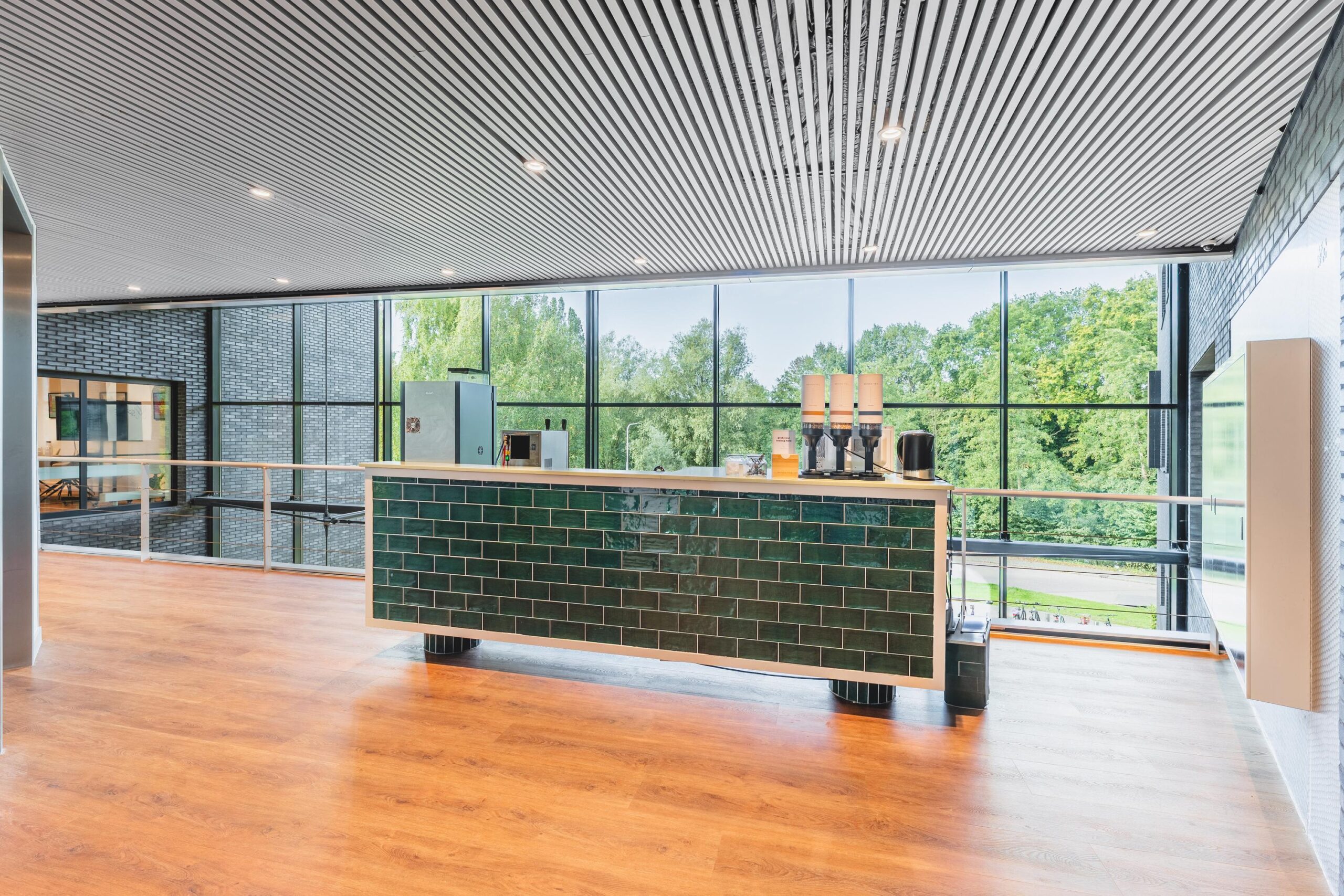 Moderne koffiecorner met donkergroene tegels en koffieapparatuur in een lichte ruimte met grote ramen en houten vloer.